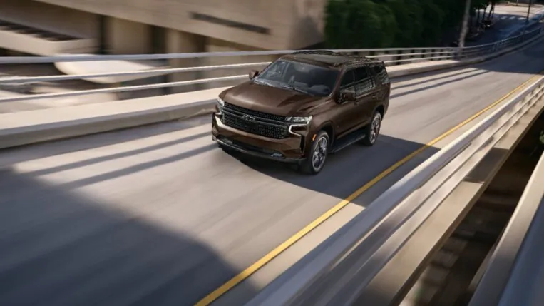 The 2023 Chevrolet Tahoe speeds along an overpass. The SUV is a metallic brown shade. We see it from an front quarter, slightly overhead angle.