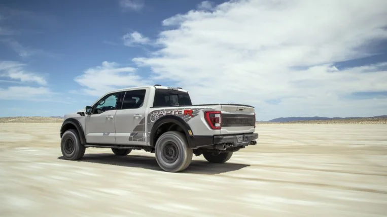 The 2023 Ford F-150 Raptor R drives through the desert. The truck is a light gray color Ford calls Azure Gray. We see it from a rear quarter angle, driving away from us to the left.