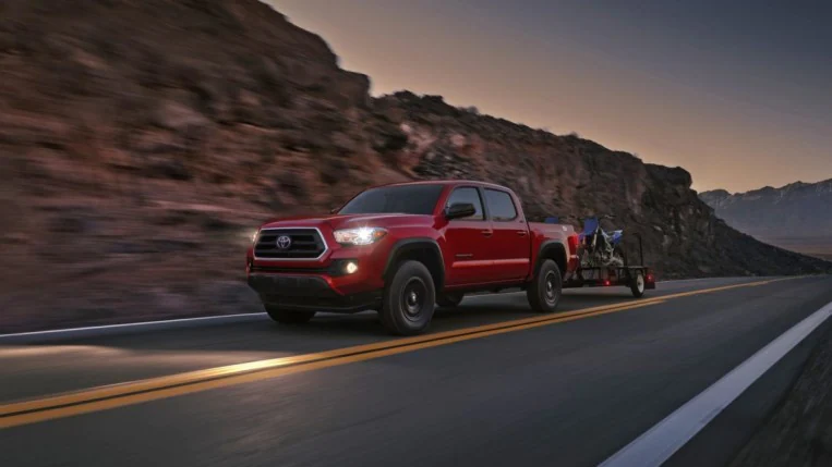 A 2023 Toyota Tacoma drives past a rocky ridge. The truck is red, and pulling a pair of motorcycles on a trailer.
