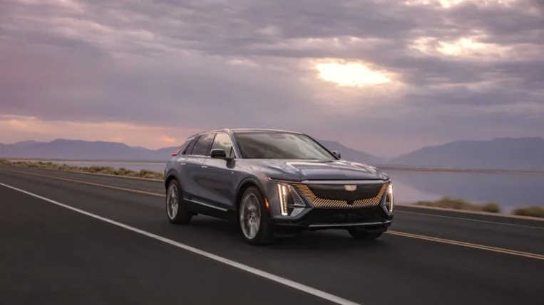 A 2024 Cadillac Lyriq electric SUV drives past a pink sunrise. The car is silver. We see it from a front quarter angle, facing slightly to our right.