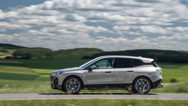 The 2023 BMW iX electric SUV rives past a green field. The car is silver. We see it in profile, facing to our left.