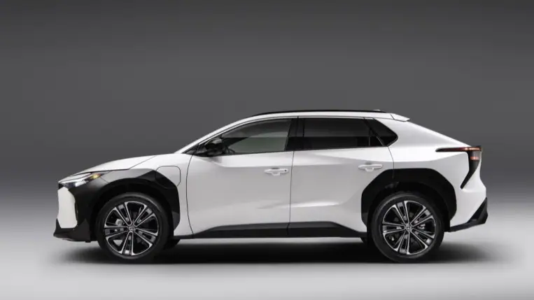 The 2023 Toyota bZ4X sits parked against a gray wall. The car is white, with thick black body cladding around the front wheels. We see it in profile. 